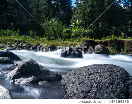 Long exposure photograph of a scenic mountain river flowing rapidly over smooth rocks through a lush green pine forest under a clear blue sky with bright white clouds. In Songgon, Banyuwangi, East Jav Long exposure photograph of a scenic mountain river flowing rapidly over smooth rocks through a lush green pine forest under a clear blue sky with bright white clouds. In Songgon, Banyuwangi, East Jav 142164586