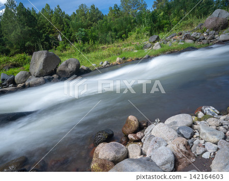 Long exposure photograph of a scenic mountain river flowing rapidly over smooth rocks through a lush green pine forest under a clear blue sky with bright white clouds. In Songgon, Banyuwangi, East Jav Long exposure photograph of a scenic mountain river flowing rapidly over smooth rocks through a lush green pine forest under a clear blue sky with bright white clouds. In Songgon, Banyuwangi, East Jav 142164603