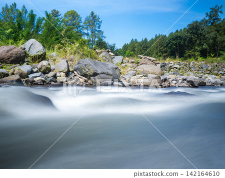 Long exposure photograph of a scenic mountain river flowing rapidly over smooth rocks through a lush green pine forest under a clear blue sky with bright white clouds. In Songgon, Banyuwangi, East Jav Long exposure photograph of a scenic mountain river flowing rapidly over smooth rocks through a lush green pine forest under a clear blue sky with bright white clouds. In Songgon, Banyuwangi, East Jav 142164610