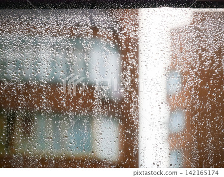 Rain drops on window glass. Abstract view of city street through wet transparent surface. Weather concept, rainy day background with blurry urban building and blurred natural light. Rain drops on window glass. Abstract view of city street through wet transparent surface. Weather concept, rainy day background with blurry urban building and blurred natural light. 142165174