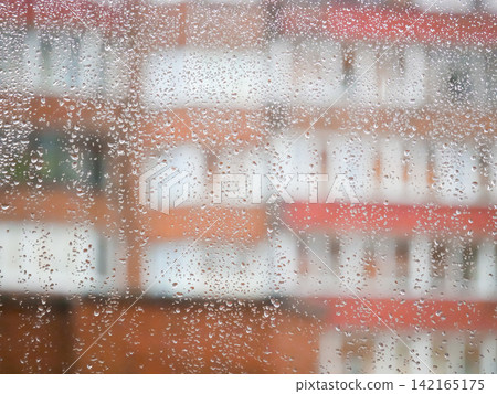 Rain drops on window glass. Abstract view of city street through wet transparent surface. Weather concept, rainy day background with blurry urban building and blurred natural light. Rain drops on window glass. Abstract view of city street through wet transparent surface. Weather concept, rainy day background with blurry urban building and blurred natural light. 142165175
