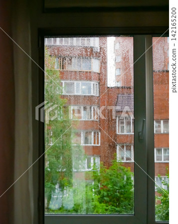 Raindrops on window glass looking out at city apartment building. Gloomy weather outside home view. Melancholy concept of staying indoor during a storm with autumn atmosphere. Raindrops on window glass looking out at city apartment building. Gloomy weather outside home view. Melancholy concept of staying indoor during a storm with autumn atmosphere. 142165200