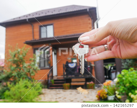 Female hand holding house key in open palm with view of wooden home patio and garden. Concept of new home ownership. Female hand holding house key in open palm with view of wooden home patio and garden. Concept of new home ownership. 142165297