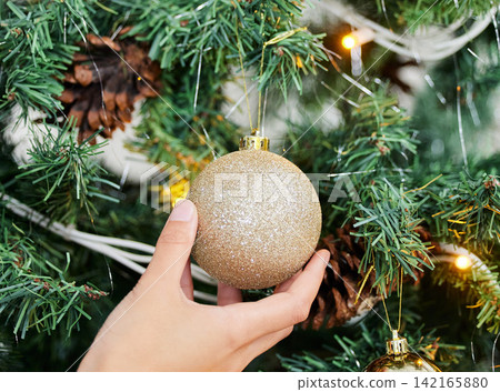 Person, hands and ball with Christmas tree for decoration, festive or holiday season at home. Closeup of ornament, lights and decor with glitter bauble in December celebration or new year preparation Person, hands and ball with Christmas tree for decoration, festive or holiday season at home. Closeup of ornament, lights and decor with glitter bauble in December celebration or new year preparation 142165880