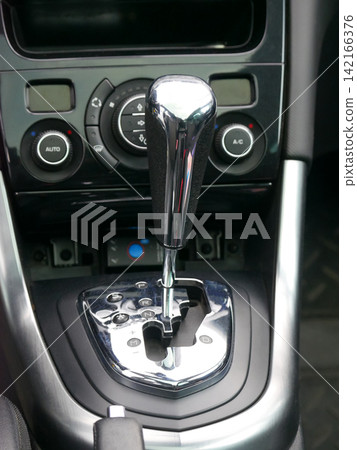 Automatic gear shifter in modern vehicle interior. Metallic transmission selector lever in car console. Driving control panel with climate settings and parking mode for automotive industry. Automatic gear shifter in modern vehicle interior. Metallic transmission selector lever in car console. Driving control panel with climate settings and parking mode for automotive industry. 142166376