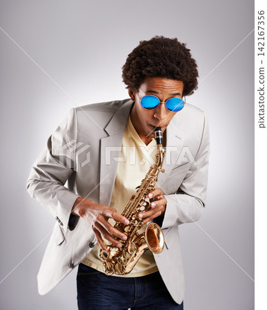 Happy, black man and music, playing the saxophone with casual suit and sunglasses in studio. African musician, jazz and instrument with confidence and trendy outfit for band with gray background Happy, black man and music, playing the saxophone with casual suit and sunglasses in studio. African musician, jazz and instrument with confidence and trendy outfit for band with gray background 142167356