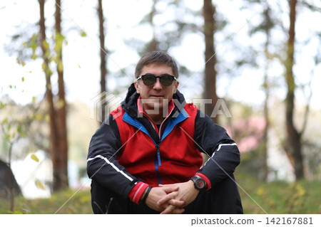 Man in sunglasses and red jacket sitting in forest. Active lifestyle, leisure activity, and connection with nature concept for outdoors. Man in sunglasses and red jacket sitting in forest. Active lifestyle, leisure activity, and connection with nature concept for outdoors. 142167881