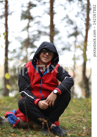 Man in sunglasses and a hooded jacket sitting on a log in a forest. Autumn outdoor activity and nature exploration concept. Man in sunglasses and a hooded jacket sitting on a log in a forest. Autumn outdoor activity and nature exploration concept. 142167882