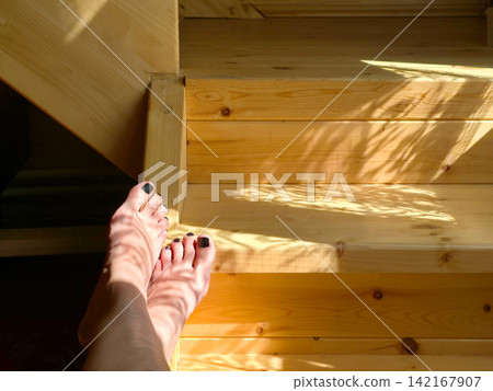 Woman feet with dark nail polish resting on wooden staircase. Sunny morning atmosphere in residential house interior. Relaxation at home, comfortable lifestyle and summer leisure concept. Woman feet with dark nail polish resting on wooden staircase. Sunny morning atmosphere in residential house interior. Relaxation at home, comfortable lifestyle and summer leisure concept. 142167907