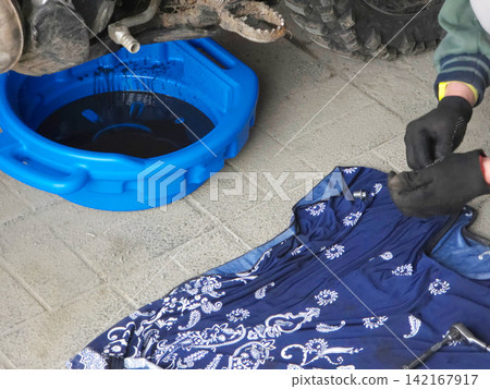 Man mechanic wearing gloves changing oil in vehicle. Automotive maintenance service with blue drain pan. Professional repairman working on dirty machine motor in garage workshop environment. Man mechanic wearing gloves changing oil in vehicle. Automotive maintenance service with blue drain pan. Professional repairman working on dirty machine motor in garage workshop environment. 142167917