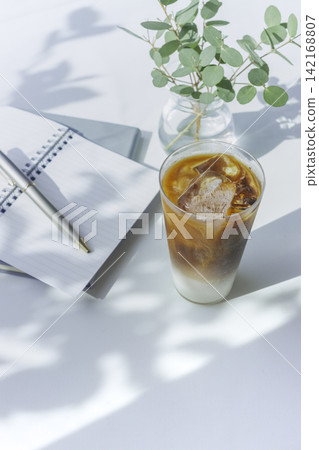 An iced cafe latte, a notebook, a pen, and eucalyptus are placed on a desk bathed in natural light. An iced cafe latte, a notebook, a pen, and eucalyptus are placed on a desk bathed in natural light. 142168807