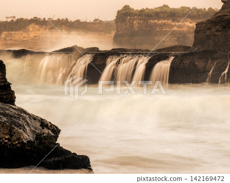 Long exposure photography of silky smooth water cascading over rocky cliff terraces at Klayar Beach (Pantai Klayar) during warm golden hour sunrise, Pacitan, East Java, Indonesia. Long exposure photography of silky smooth water cascading over rocky cliff terraces at Klayar Beach (Pantai Klayar) during warm golden hour sunrise, Pacitan, East Java, Indonesia. 142169472