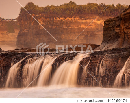 Long exposure photography of silky smooth water cascading over rocky cliff terraces at Klayar Beach (Pantai Klayar) during warm golden hour sunrise, Pacitan, East Java, Indonesia. Long exposure photography of silky smooth water cascading over rocky cliff terraces at Klayar Beach (Pantai Klayar) during warm golden hour sunrise, Pacitan, East Java, Indonesia. 142169474