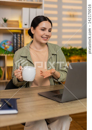 Young woman on home table making video call on laptop and drinking coffee during remote work session Young woman on home table making video call on laptop and drinking coffee during remote work session 142170016