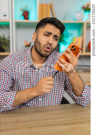 Indian man on home table trying broken charger on smartphone feeling upset about charging port issue Indian man on home table trying broken charger on smartphone feeling upset about charging port issue 142170020