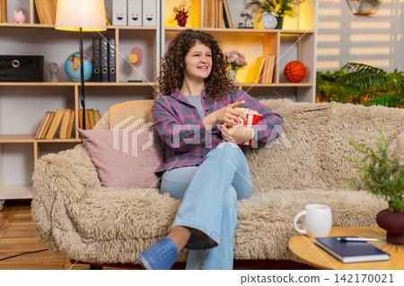Young woman on home sofa watching fun film with popcorn laughing in surprise during home movie night Young woman on home sofa watching fun film with popcorn laughing in surprise during home movie night 142170021