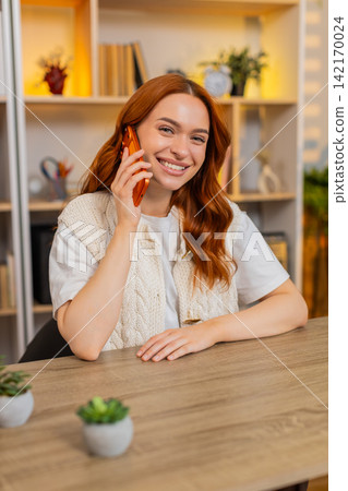 Young woman on home table talking on smartphone with expressive reaction during phone call indoors Young woman on home table talking on smartphone with expressive reaction during phone call indoors 142170024