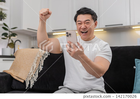 Asian man celebrates app win with smartphone shares good news via social media chat smiling excited Asian man celebrates app win with smartphone shares good news via social media chat smiling excited 142170026