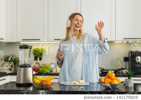 Young woman slicing banana for smoothie blender while dancing with headphones for healthy fit diet Young woman slicing banana for smoothie blender while dancing with headphones for healthy fit diet 142170029