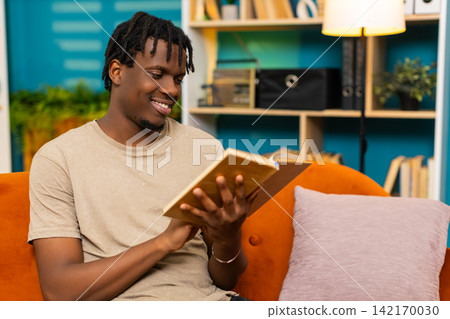 African American young man on home sofa reading book turning page with relaxed smile during leisure African American young man on home sofa reading book turning page with relaxed smile during leisure 142170030