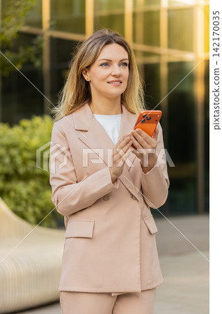 Young woman manager walking in city street typing message on smartphone near modern business center Young woman manager walking in city street typing message on smartphone near modern business center 142170035