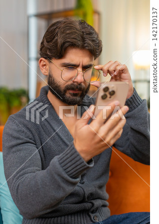 Indian man with eyeglasses squints at smartphone screen tries to read small text poor vision focus Indian man with eyeglasses squints at smartphone screen tries to read small text poor vision focus 142170137