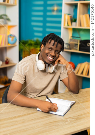 African American young man on home table writing notes in notebook with pen for study task planning 142170140