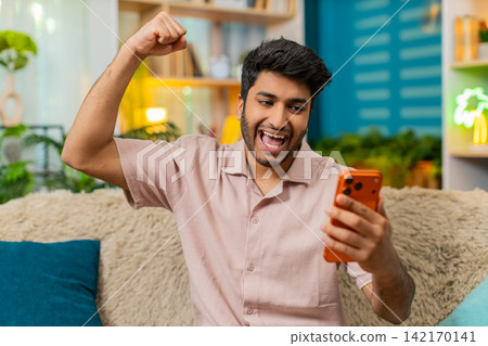 Indian man on home sofa celebrating online game win with smartphone good news victory gesture happy Indian man on home sofa celebrating online game win with smartphone good news victory gesture happy 142170141