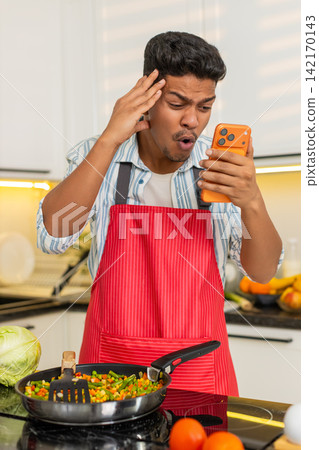 Indian man reading bad smartphone message while frying vegetables in kitchen for healthy meal prep 142170143