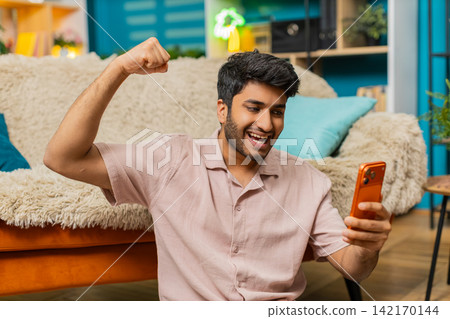 Indian man on home floor celebrating smartphone win with happy smile excited victory gesture indoors Indian man on home floor celebrating smartphone win with happy smile excited victory gesture indoors 142170144