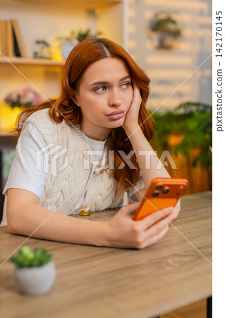Young woman on home table scrolling smartphone news feed with bored expression in living room home Young woman on home table scrolling smartphone news feed with bored expression in living room home 142170145