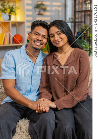Indian couple man and woman sitting on home sofa smiling hugging and sharing loving portrait at home Indian couple man and woman sitting on home sofa smiling hugging and sharing loving portrait at home 142170151