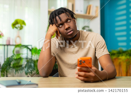 African American young man on home table bored with smartphone scrolling social media news indoors African American young man on home table bored with smartphone scrolling social media news indoors 142170155