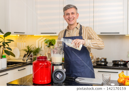 Middle-aged man pouring milk into blender with banana for protein shake meal prep in kitchen at home Middle-aged man pouring milk into blender with banana for protein shake meal prep in kitchen at home 142170159