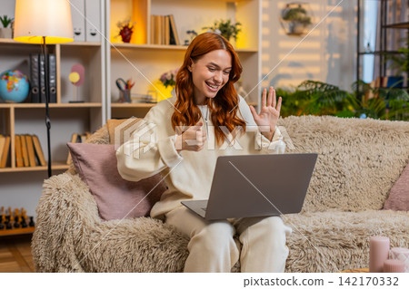 Young woman on home sofa greeting during laptop video call and speaking on friendly online meeting Young woman on home sofa greeting during laptop video call and speaking on friendly online meeting 142170332