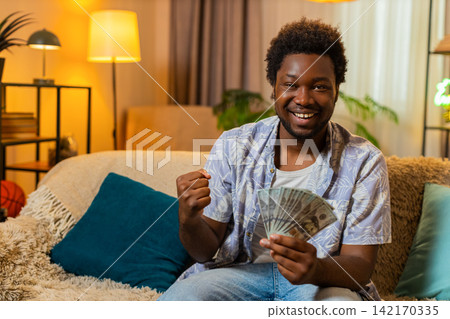 African American young man on home sofa counts cash and plans purchase with satisfied smile indoors African American young man on home sofa counts cash and plans purchase with satisfied smile indoors 142170335