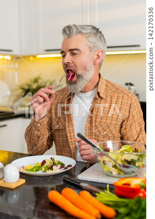 Middle-aged man enjoying fresh vegetable salad in home kitchen for healthy diet nutrition lifestyle 142170336