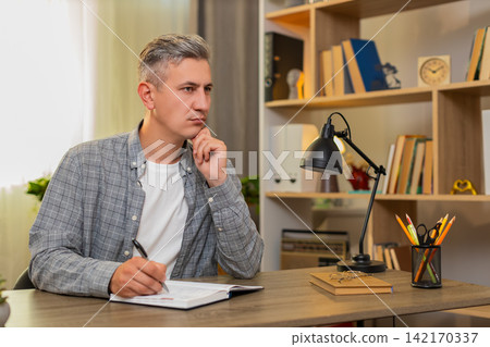 Middle-aged man on home table plans weekly tasks in calendar notebook and marks done goals clearly Middle-aged man on home table plans weekly tasks in calendar notebook and marks done goals clearly 142170337
