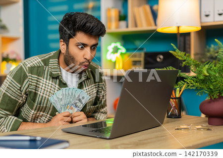 Indian man on home table holding cash and celebrating online game win while looking at laptop screen Indian man on home table holding cash and celebrating online game win while looking at laptop screen 142170339