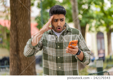 Indian man reading bad news walking in city street checking smartphone with shocked upset reaction Indian man reading bad news walking in city street checking smartphone with shocked upset reaction 142170343