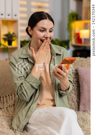Young woman on home sofa celebrating smartphone win with joyful surprise in cozy living room indoors Young woman on home sofa celebrating smartphone win with joyful surprise in cozy living room indoors 142170344
