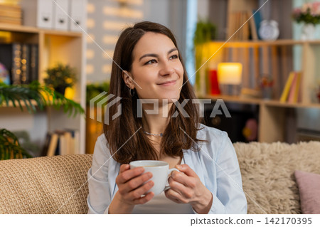 Young woman on home sofa enjoying coffee and thinking about future plans with gentle smile indoors 142170395