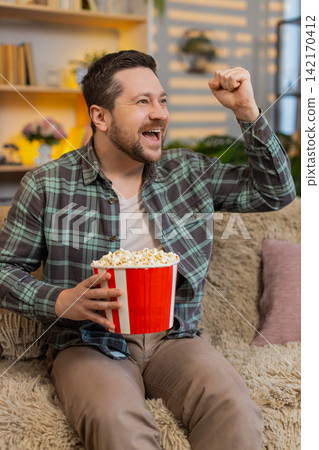 Young man on home sofa cheering with popcorn while watching sports victory and celebrating success Young man on home sofa cheering with popcorn while watching sports victory and celebrating success 142170412