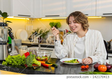 Young woman enjoying fresh vegetable salads in home kitchen smiling during healthy diet meal plans 142170413
