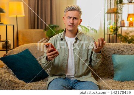 Middle-aged man on home sofa upset after smartphone money charge and online game loss with bad news 142170439