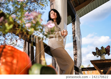 Woman in cozy sweater holds mug on veranda, calm gazing at autumn sky. Fall atmosphere, autumn cozy lifestyle, wellness Woman in cozy sweater holds mug on veranda, calm gazing at autumn sky. Fall atmosphere, autumn cozy lifestyle, wellness 142170622
