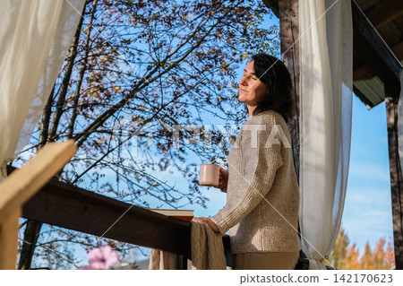 Woman with eyes closed enjoying autumn sun on cozy wooden porch with mug in hand. Mindfulness, mental health, wellness, country lifestyle Woman with eyes closed enjoying autumn sun on cozy wooden porch with mug in hand. Mindfulness, mental health, wellness, country lifestyle 142170623