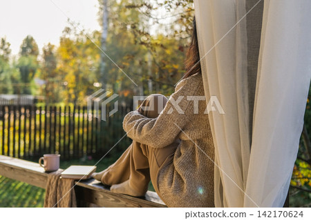 Woman sitting on cozy terrace railing in autumn, looking at garden views with book and mug nearby. Great for mental health, cozy lifestyle and wellness themes Woman sitting on cozy terrace railing in autumn, looking at garden views with book and mug nearby. Great for mental health, cozy lifestyle and wellness themes 142170624