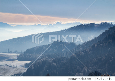 Central Eastern Alps near Micheldorf, Carinthia, Austria. A winter landscape showing a foggy valley with snow-covered mountains under a colorful sunrise sky. Central Eastern Alps near Micheldorf, Carinthia, Austria. A winter landscape showing a foggy valley with snow-covered mountains under a colorful sunrise sky. 142170810