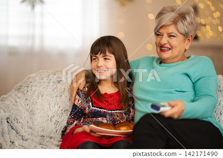 Grandmother granddaughter watching TV together laughing eating snacks Grandmother granddaughter watching TV together laughing eating snacks 142171490